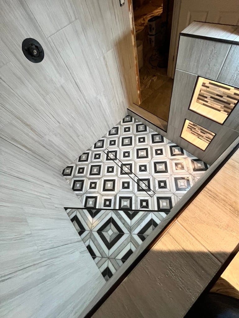 Black and white geometric mosaic shower floor with gray wood-look walls, mid-install