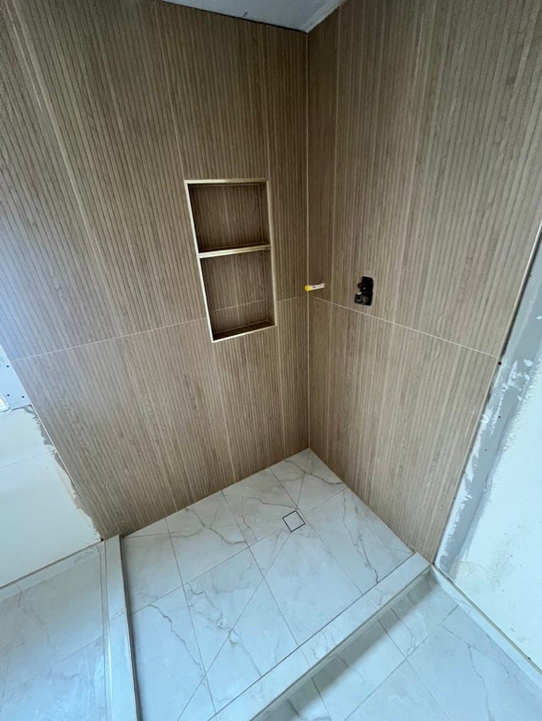 Shower walls in wood-look slat porcelain mid-install with a recessed niche and marble-look floor
