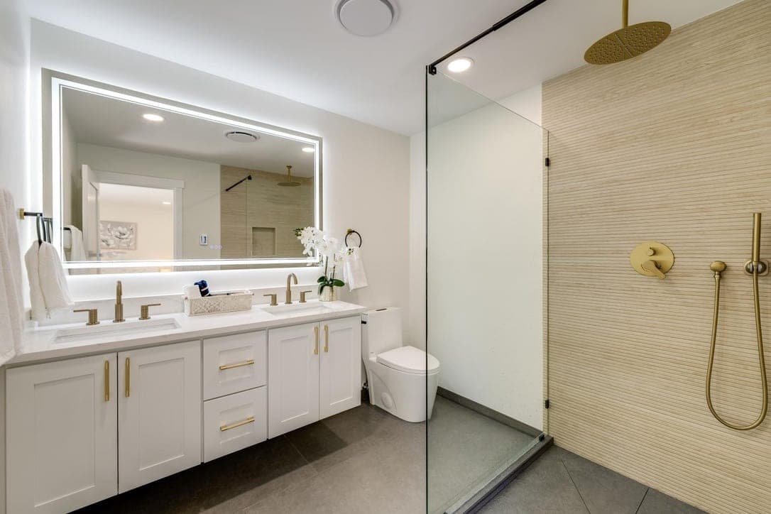 Double vanity with backlit mirror and brass faucets next to the slat tile shower